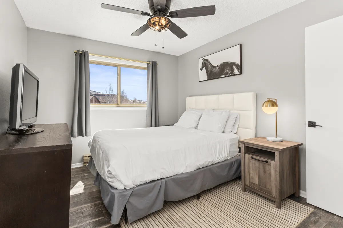 A serene queen bedroom featuring a plush bed with white bedding, a wooden nightstand, and a television. Natural light streams through a window, creating a tranquil atmosphere ideal for restful nights.