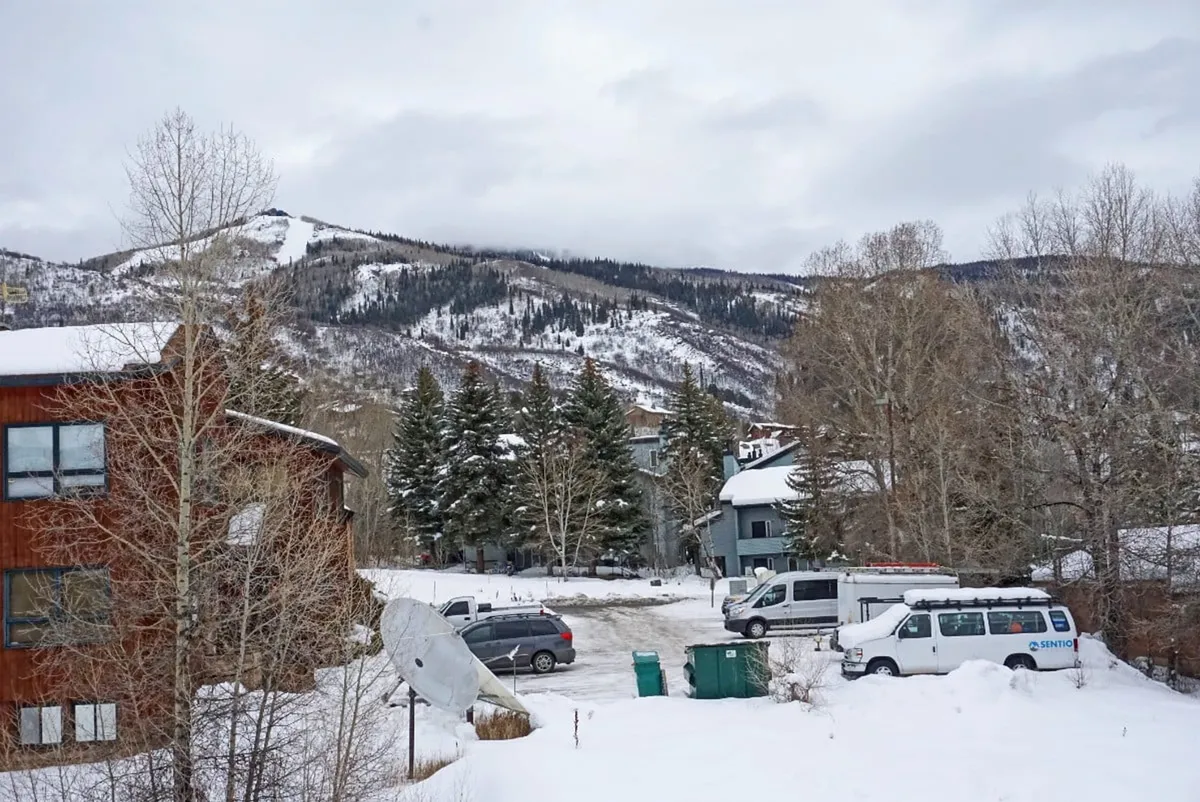 A wide exterior view capturing snow-covered mountains, evergreen trees, and several buildings nestled at the base. The landscape evokes the excitement of winter sports, with the buildings suggesting proximity to essential resort amenities and gondola access.