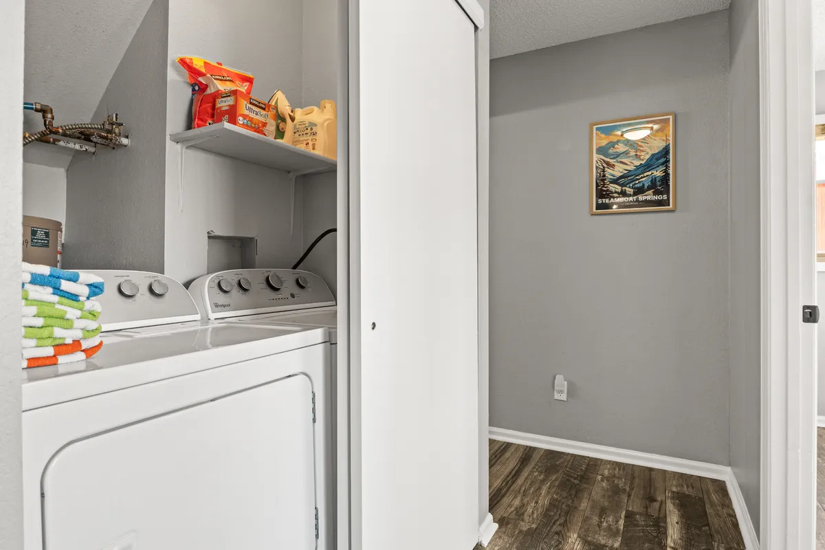 A tidy laundry nook featuring a front-loading washing machine, shelves stocked with laundry essentials, and mountain-themed artwork adding a personal touch. The space is efficient and conveniently located for daily routines or longer stays.