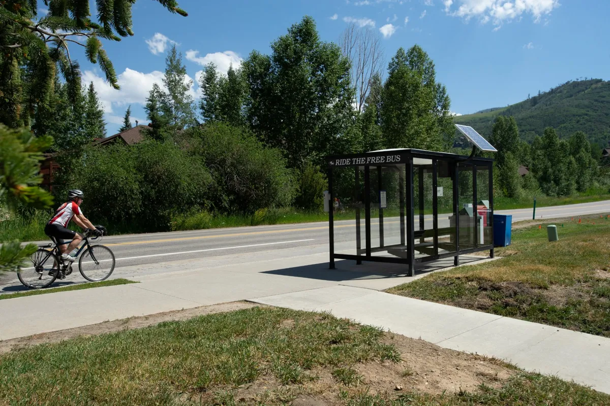 A vibrant exterior scene with a cyclist riding past a bus stop set against a backdrop of greenery and distant mountains. The bus stop emphasizes easy access to local transportation and the active, family-friendly neighborhood vibe.