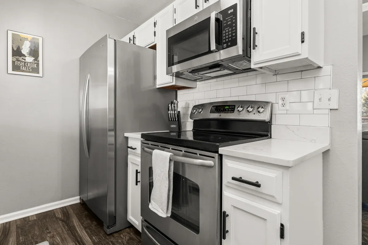 A bright, modern kitchen featuring white cabinetry, gleaming stainless steel appliances, and a spacious marble-like countertop. The layout is open and inviting, designed for both cooking and socializing. Natural light fills the room, highlighting the ample prep space and clean, contemporary finishes.