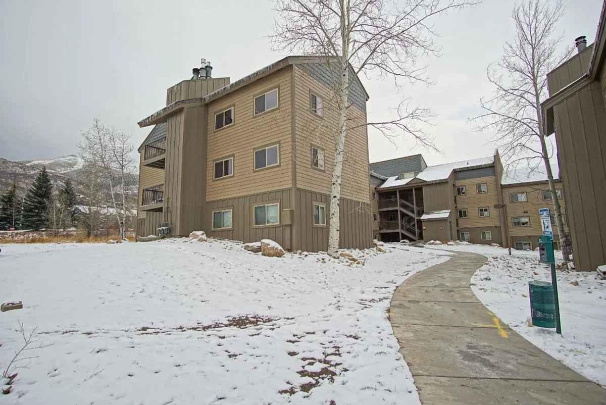 A multi-story building stands amid a snowy landscape, with a pathway leading up to the entry. Evergreen trees and soft white snow enhance the tranquil ambiance, evoking a peaceful mountain retreat.