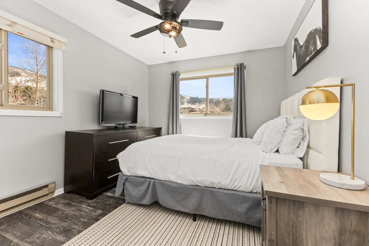 A cozy, modern bedroom with a bed, window, television, nightstand, and artwork. The room exudes a warm ambiance with mountain views visible through the window, inviting relaxation and leisure.
