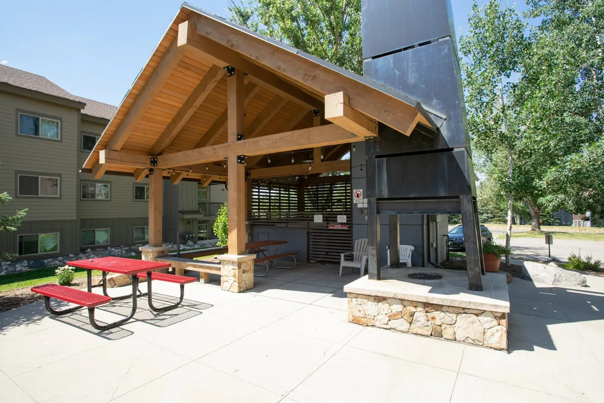 An outdoor gathering space featuring a sturdy wooden structure, stone seating area, and picnic tables. The scene evokes scenes of barbecuing, friendly conversation, and relaxed outdoor meals.