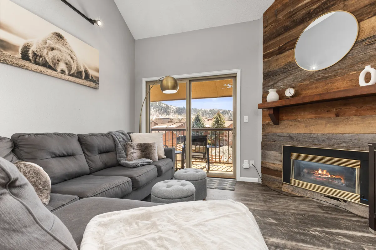A warm and welcoming living room with a large sectional sofa arranged around a striking reclaimed barn-wood fireplace. A spacious wood accent wall and large window with views create a sense of openness and connection to the outdoors, while soft lighting and plush textiles invite guests to relax and socialize.