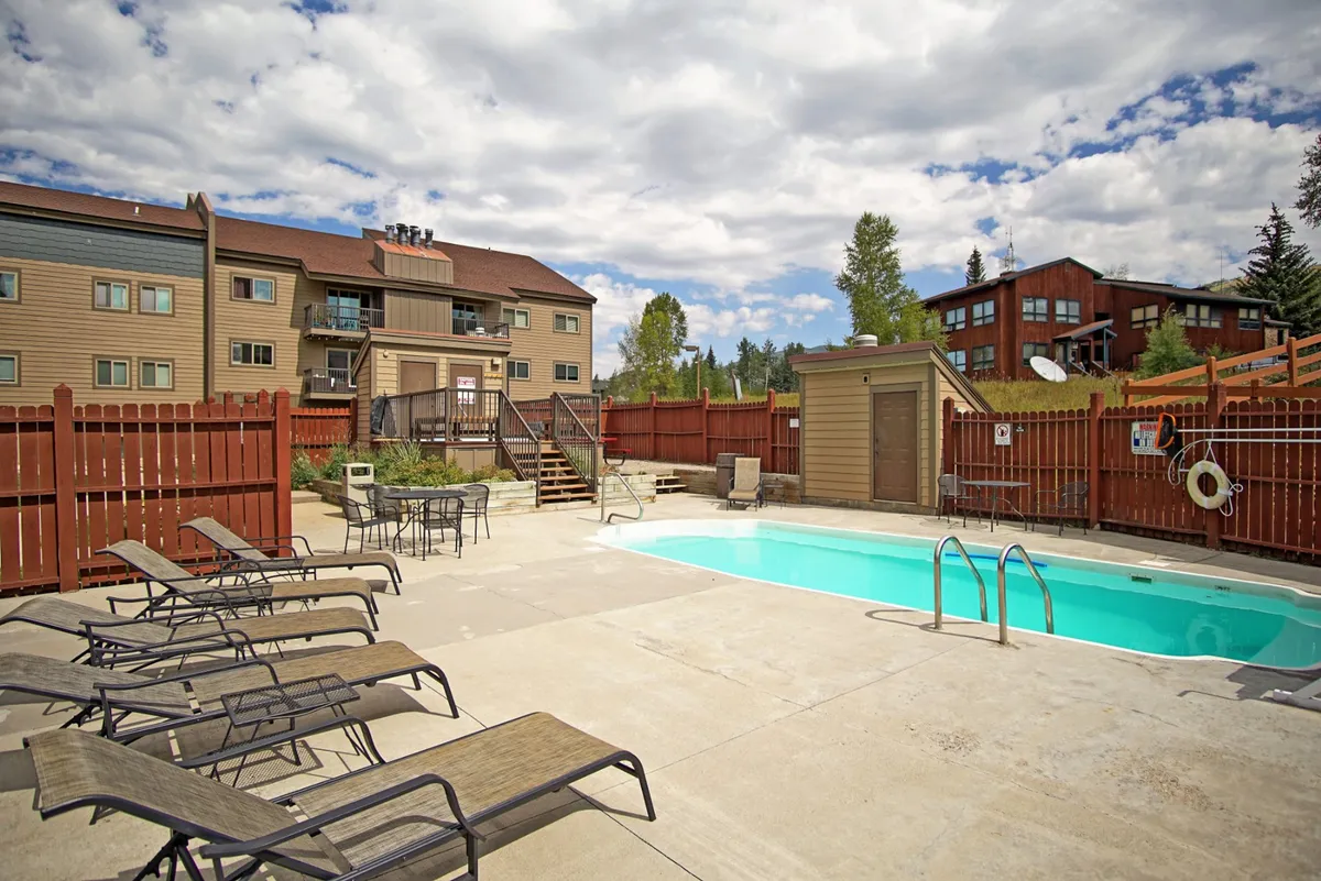 This image features a sparkling swimming pool surrounded by comfortable lounge chairs, a wooden fence, and glimpses of the property's buildings in the background. The pool area is bathed in natural light, inviting guests to swim or relax in the sun.