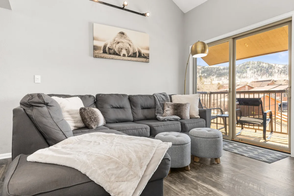 An inviting sectional sofa faces a large window that opens to a balcony view, with playful bear artwork adding a touch of whimsy. The layout suggests a perfect spot for movie nights or morning coffee with glimpses of the surrounding mountains.