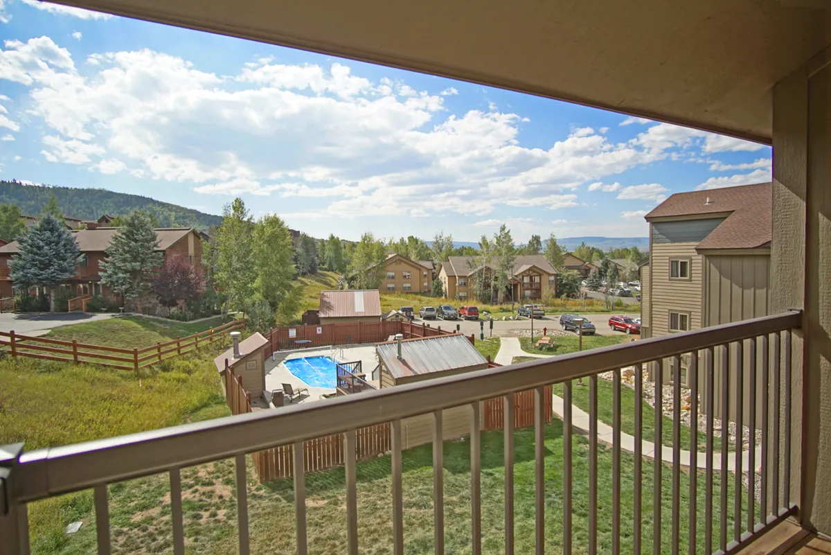 A view from the condo's balcony overlooking the on-site swimming pool surrounded by lush greenery, with other buildings in the development visible nearby. The scene is tranquil, hinting at summer enjoyment and community gathering spaces.