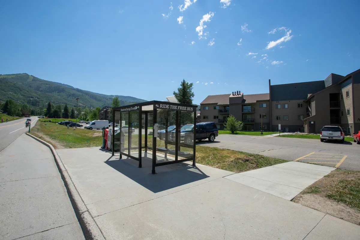 This image shows a bus stop shelter set against a backdrop of surrounding mountains and an apartment building. The area is casual and relaxed, with a pathway for walking or biking and an open view of the mountainous landscape.
