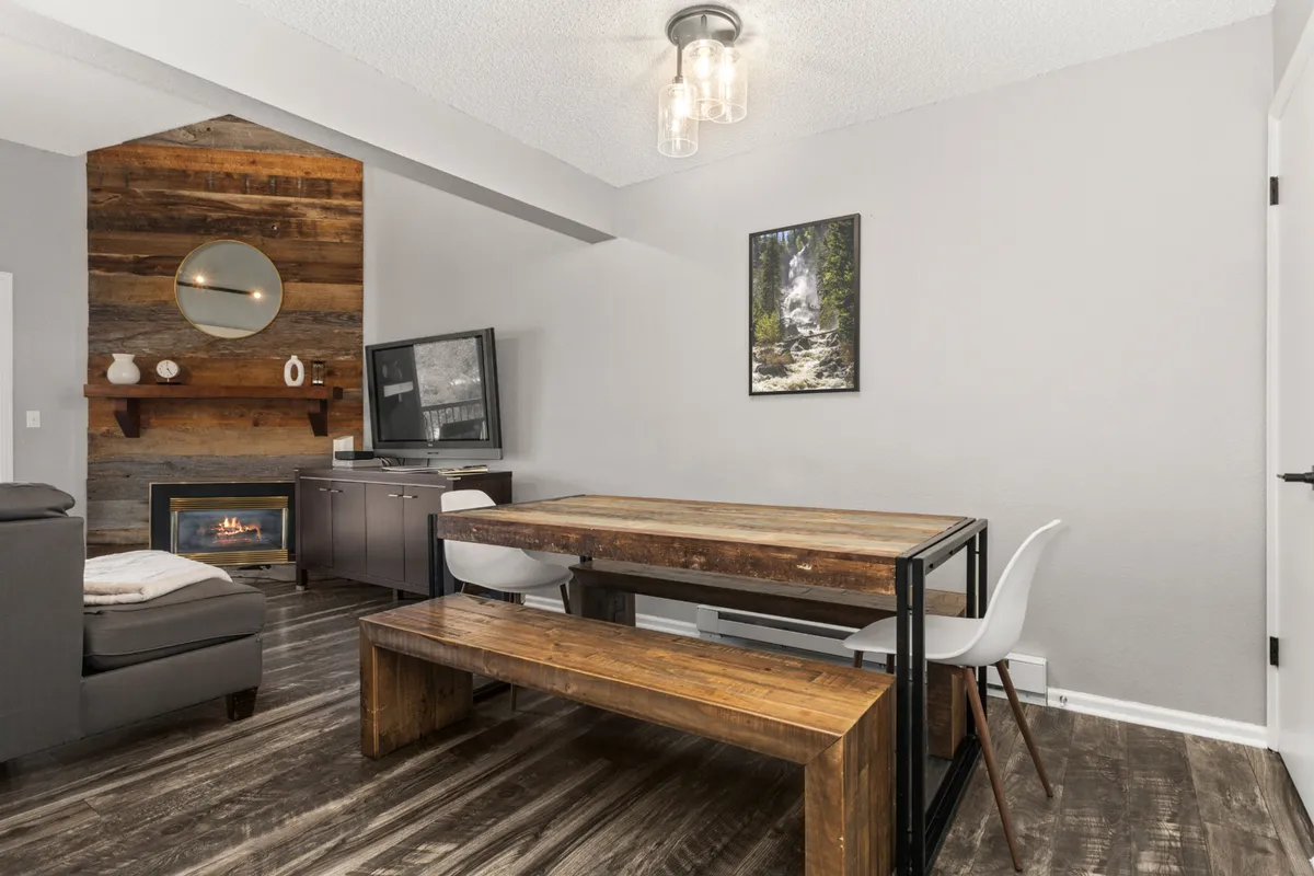 A bright, open-concept space that merges the dining area and living room. The wooden dining table sits adjacent to the fireplace and a wall-mounted TV, with rustic touches like a wooden wall feature and framed artwork enhancing the stylish mountain ambiance. The layout encourages flow and conversation.